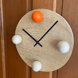 Puede incluir: Un reloj redondo de madera con manecillas negras y tres pomos de madera blancos y un pomo naranja.