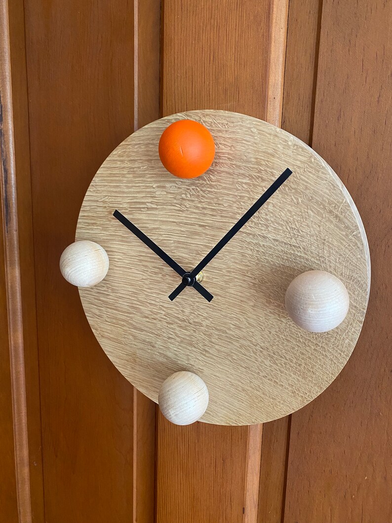 Puede incluir: Un reloj redondo de madera con manecillas negras y tres pomos de madera blancos y un pomo naranja.