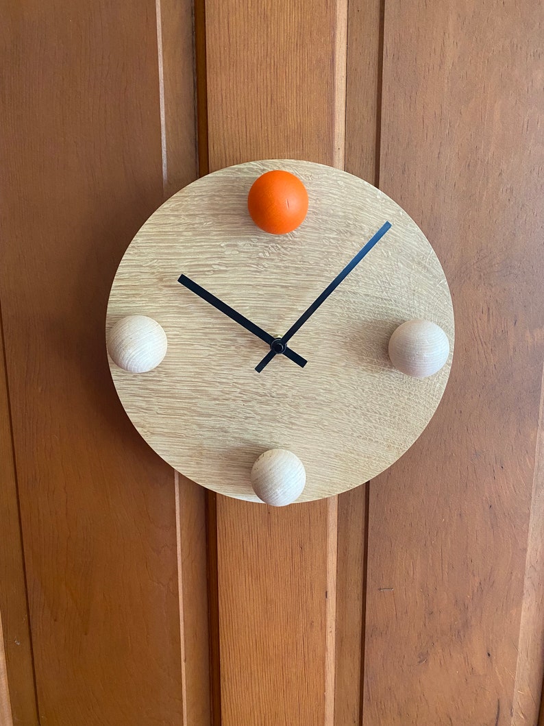 Puede incluir: Un reloj redondo de madera con manecillas negras y tres pomos de madera. El reloj tiene un pomo naranja en la parte superior.