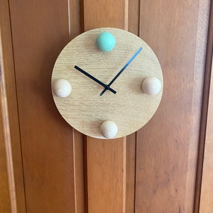 Puede incluir: Un reloj redondo de madera con manecillas negras y tres pomos de madera. Los pomos est&aacute;n en las posiciones de las 12, las 3 y las 9. El pomo de las 12 est&aacute; pintado de color verde claro.