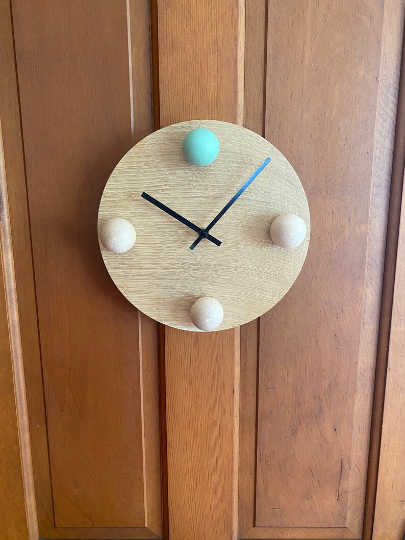 Puede incluir: Un reloj redondo de madera con manecillas negras y tres pomos de madera. Los pomos est&aacute;n en las posiciones de las 12, las 3 y las 9. El pomo de las 12 est&aacute; pintado de color verde claro.