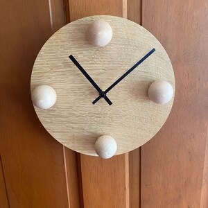 Puede incluir: Un reloj de madera redondo con manecillas negras y cuatro pomos de madera alrededor del borde.