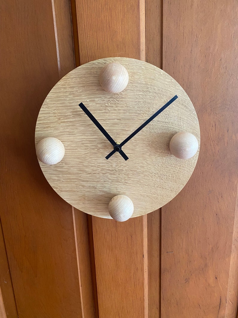 Puede incluir: Un reloj de madera redondo con manecillas negras y cuatro pomos de madera alrededor del borde.