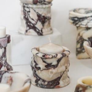 Puede incluir: Una colecci&oacute;n de objetos decorativos de piedra tallada con un patr&oacute;n de m&aacute;rmol en color crema y morado oscuro. El conjunto incluye varios portavelas, un tarro con tapa y cuencos peque&ntilde;os, cada uno con vetas &uacute;nicas y pulidas.