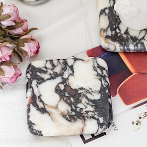 May include: A square marble coaster featuring a polished white surface with prominent black and dark brown veining. The coaster has rounded corners and a smooth, glossy finish, displaying a natural stone pattern throughout the piece.