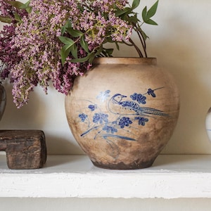 May include: Three ceramic vessels of varying sizes and shapes. The left vessel is grey with blue floral patterns, the center vessel is tan with a blue bird and floral design, and the right vessel is white with blue floral motifs. Purple flowers are inside.