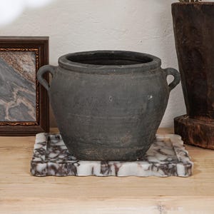 May include: A dark gray, aged-looking ceramic pot with two handles sits on a marble tray with a decorative edge. The tray has a white and gray marbled pattern. A framed picture and a wooden vase with white flowers are in the background.