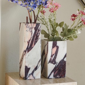 May include: Two rectangular marble vases with white, dark purple, and soft pink veining. The taller vase holds a cluster of blue flowers, while the shorter vase contains a mix of pink and white flowers with green leaves and stems.