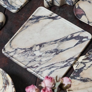May include: A collection of polished marble trays and dishes featuring a cream-colored base with prominent dark purple and black veining. The set includes a square tray, an oval dish, and a scalloped-edge piece, all displaying natural stone patterns.