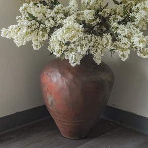 May include: A large, dark brown, textured clay vase with a wide, rounded body and a tapered base, filled with a dense arrangement of white flowering branches featuring small, delicate blossoms and dark green leaves.
