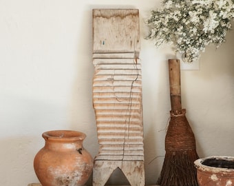 Puede incluir: Una tabla de lavar de madera desgastada se encuentra en posición vertical junto a una pequeña maceta de terracota, una escoba rústica con mango de madera y un cuenco ancho de terracota. Estos objetos están dispuestos sobre una superficie de madera con un ramo de flores blancas.