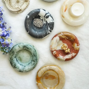 May include: A collection of decorative stone dishes in various colors and patterns. Some dishes hold small items like a candle, a flower, and jewelry. The dishes are round with a central indentation.