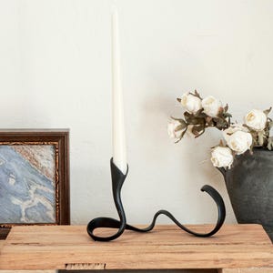 May include: A black metal candle holder with a curved design, holding a white taper candle. The candle holder sits on a wooden platform. A framed picture and a vase of white flowers are in the background.