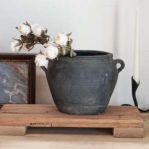 May include: A dark gray, rustic ceramic pot with two handles, holding white roses. The pot sits on a wooden platform. A framed picture and a tall white candle in a black holder are in the background.