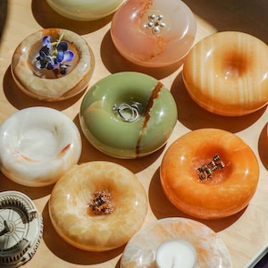 May include: A collection of smooth, donut-shaped stone trinket dishes in various colors including pale green, orange, and white. Some dishes hold small items like rings, a tea light candle, or a small blue flower, while others remain empty.