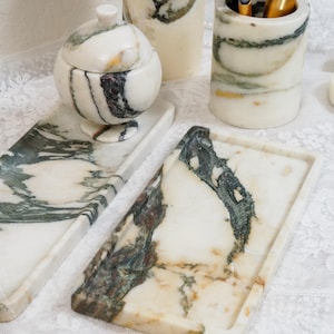 May include: A collection of white marble bathroom accessories featuring green and brown veining. The set includes two rectangular trays, a lidded spherical container, a tall cylindrical canister, and a cup holding three makeup brushes.