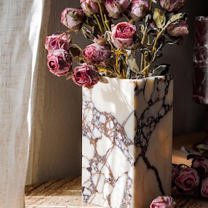 May include: A rectangular white marble vase with prominent dark brown and grey veining, filled with a bouquet of dried dusty pink roses. The vase has a smooth, polished surface and stands upright, displaying a natural stone pattern throughout its structure.