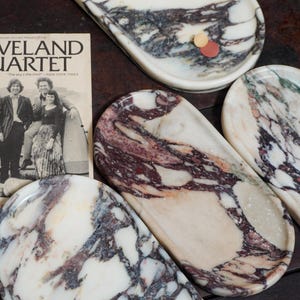 May include: Three oval marble trays featuring white, cream, and deep purple veining patterns. A vintage paper advertisement for The Cleveland Quartet with the text "The sky's the limit!" is positioned next to the trays on a dark surface.