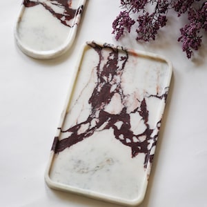 May include: Two white marble trays with deep burgundy veining. One tray is rectangular with rounded corners and raised edges, while the other is oval-shaped. The natural stone surfaces feature intricate, dark red patterns against a white base.