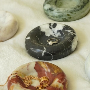 May include: A collection of handcrafted stone ring dishes in various colors and patterns. One dish holds a gold ring with a diamond, and another holds a gold necklace and ring. The dishes are made of marble and granite.