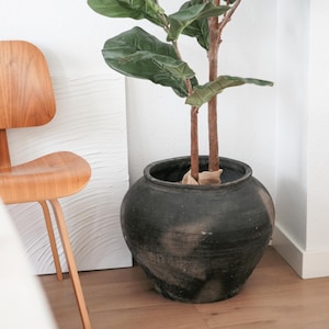 May include: A large, dark gray ceramic pot holds a faux fiddle-leaf fig tree with large, green leaves and brown stems. The pot sits on a light brown hardwood floor, near a wooden chair and a white wall.