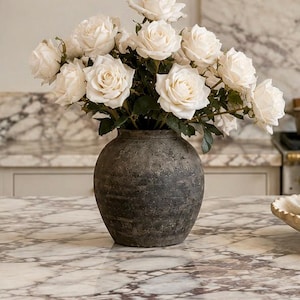 May include: A dark gray, textured vase filled with a bouquet of white roses. The vase sits on a marble countertop. The roses are in full bloom, with many petals visible.