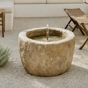 May include: An outdoor patio scene featuring a stone fountain with a water spout. Beige cushions adorn a built-in bench, complemented by a wooden table and woven chair. Large clay pots and lush greenery add to the serene ambiance.