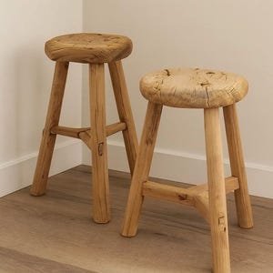 May include: Two rustic wooden stools with natural wood grain. Each stool has a round seat and three sturdy legs connected by a lower support. The stools are made of light-colored wood and are placed on a wooden floor.
