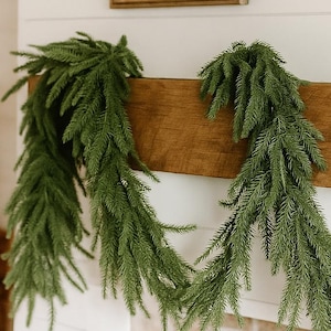 May include: A green faux fir garland draped over a wooden mantel. The garland is arranged in a flowing pattern, adding a touch of nature to the interior. A wooden framed mirror and a woven wall decor are visible in the background.
