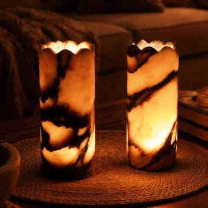 May include: Two illuminated, cylindrical lamps with scalloped edges. The lamps are made of a light-colored material with dark brown veining. They sit on a round, woven mat on a wooden table, creating a warm, inviting ambiance.