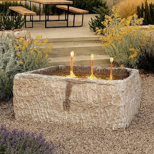 May include: A stone fountain with three flame-like water features. The fountain is set in a gravel area, with a wooden table and benches in the background. Landscaping includes yellow and purple plants.