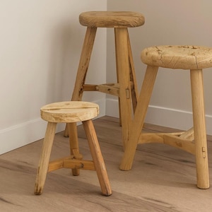 May include: Three wooden stools of varying heights. The stools have round seats and three legs. The wood has a natural, light brown color. The stools are placed in a corner of a room with light-colored walls and a wooden floor.