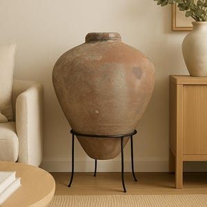 May include: A large, antique-style terracotta vase with a wide body and narrow neck, displayed on a black metal stand. The vase has a weathered, earthy appearance, and the stand has a simple, minimalist design. The vase is a decorative home decor item.