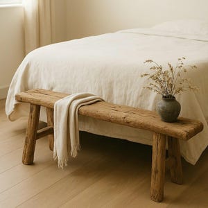 May include: A rustic wooden bench with a weathered texture, featuring four sturdy legs and a rectangular seat. A cream-colored textile throw is draped over the bench, and a dark gray ceramic vase containing dried flowers sits on the right side.