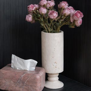 Puede incluir: Una composici&oacute;n decorativa con una caja de pa&ntilde;uelos de m&aacute;rmol rosa, un jarr&oacute;n crema lleno de rosas rosas y un libro. Los objetos se colocan sobre un fondo oscuro y texturizado, creando una escena elegante.