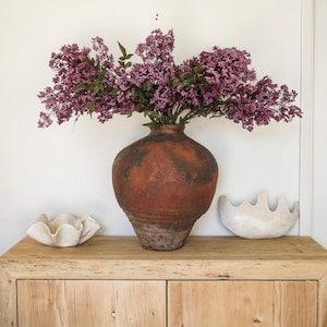 May include: A large, round, terracotta-colored clay vase with a weathered, dark-toned finish, filled with a dense arrangement of purple lilac flowers. Two small, white, wavy-edged ceramic bowls sit on either side of the vase on a wooden surface.