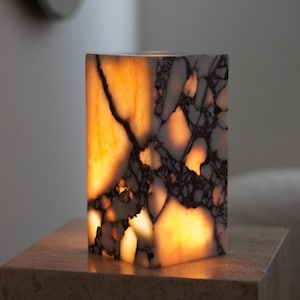 May include: A rectangular stone lamp with a polished surface featuring a white and dark grey marbled pattern. The translucent material is illuminated from within, casting a warm, glowing orange light through the natural veins of the stone.