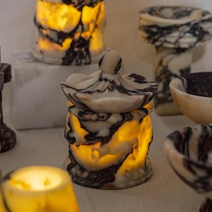 May include: A collection of translucent stone containers with lids, featuring a marbled pattern of white, cream, and dark brown veins. Each vessel is illuminated from within, casting a warm, soft glow that highlights the natural veining of the stone.