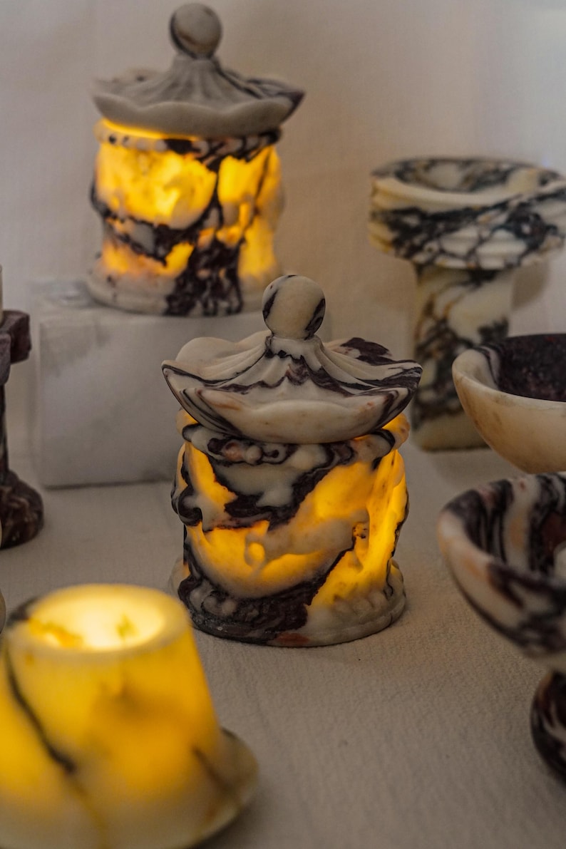 Puede incluir: Una colecci&oacute;n de recipientes de piedra transl&uacute;cida con tapa, con un patr&oacute;n veteado en blanco, crema y marr&oacute;n oscuro. Cada recipiente est&aacute; iluminado desde el interior, proyectando un brillo c&aacute;lido que resalta las vetas naturales de la piedra.
