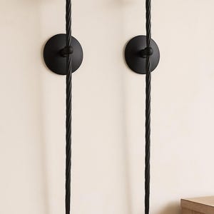 May include: Two black metal wall sconces. Each sconce features a circular base, a central sphere, and a long, twisted stem that tapers to a point. The top has a small, flat disc to hold a candle. The sconces are mounted on a light-colored wall.