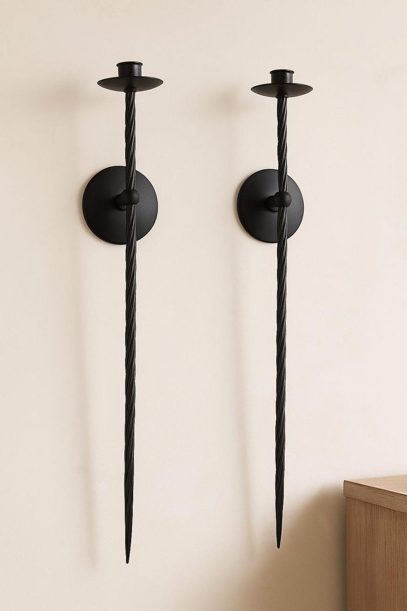 May include: Two black metal wall sconces. Each sconce features a circular base, a central sphere, and a long, twisted stem that tapers to a point. The top has a small, flat disc to hold a candle. The sconces are mounted on a light-colored wall.