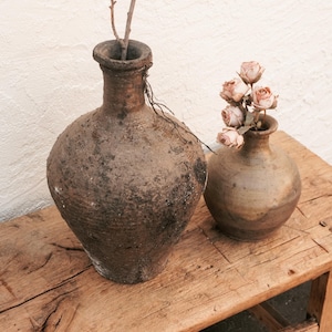 May include: Two rustic, earthenware vases sit on a weathered wooden bench. The larger vase is dark brown with a textured surface and holds a twig. The smaller vase, also brown, contains dried pink roses. Home decor.