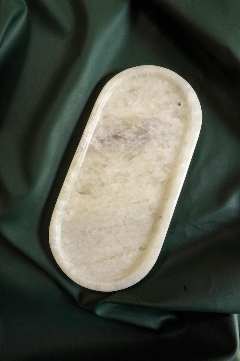 May include: A light green, oval-shaped tray with a polished surface. The tray has a slightly textured appearance with subtle variations in color. A small hole is visible near one end. The tray is set against a dark green background.