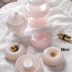 May include: A collection of pale pink stone objects including a tiered pedestal, a lidded sphere, and four ring-shaped dishes. One dish holds a gold-toned necklace with pearls, one holds a white tea light candle, and one holds a gold ring. Text reads Mini and Small.