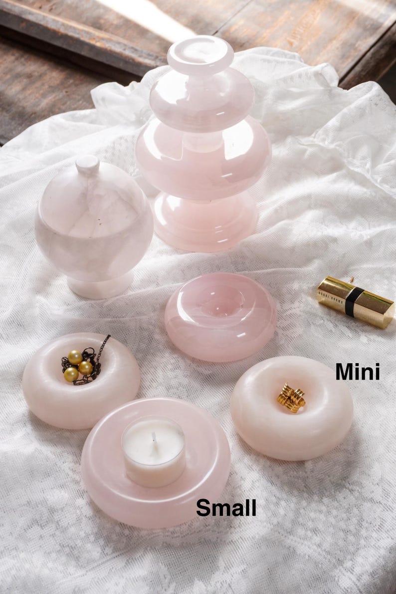 May include: A collection of pale pink stone objects including a tiered pedestal, a lidded sphere, and four ring-shaped dishes. One dish holds a gold-toned necklace with pearls, one holds a white tea light candle, and one holds a gold ring. Text reads Mini and Small.