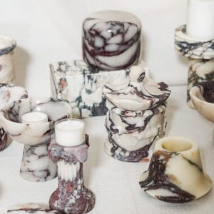 Puede incluir: Una colecci&oacute;n de objetos decorativos hechos de piedra veteada en blanco, crema y morado oscuro. El conjunto incluye varios cuencos, portavelas, un tarro con tapa y una caja rectangular, todos con patrones de remolinos &uacute;nicos y superficies pulidas.