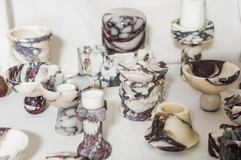 Puede incluir: Una colecci&oacute;n de objetos decorativos hechos de piedra veteada en blanco, crema y morado oscuro. El conjunto incluye varios cuencos, portavelas, un tarro con tapa y una caja rectangular, todos con patrones de remolinos &uacute;nicos y superficies pulidas.