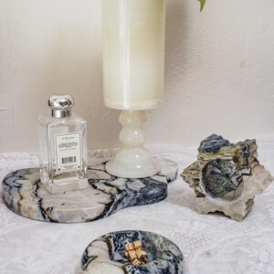 Puede incluir: Colecci&oacute;n de objetos decorativos de piedra, que incluye una bandeja de m&aacute;rmol con un frasco de perfume, un candelabro y una escultura de piedra tallada. Los objetos est&aacute;n dispuestos sobre un mantel de encaje blanco.