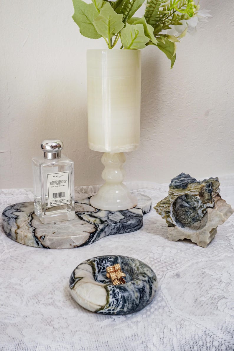 Puede incluir: Colecci&oacute;n de objetos decorativos de piedra, que incluye una bandeja de m&aacute;rmol con un frasco de perfume, un candelabro y una escultura de piedra tallada. Los objetos est&aacute;n dispuestos sobre un mantel de encaje blanco.