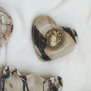 May include: An oval-shaped cameo brooch featuring a profile portrait in cream and brown tones, set within an ornate, antique-gold colored metal frame decorated with small floral accents and a bow detail at the top, resting on a heart-shaped stone.